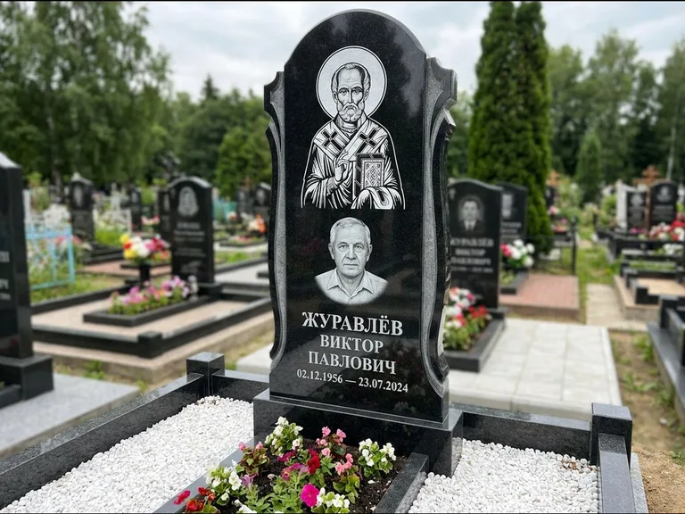 Памятник с гравировкой иконы: стела с образом в полный рост на обороте — гравировка образа Матроны Московской, низкие кусты спиреи в цвету, работа «Лебедева» в Барнауле